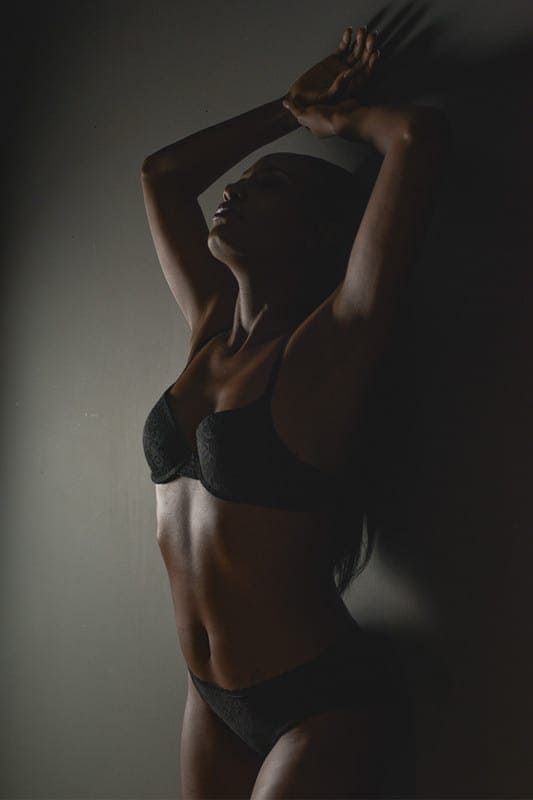 Boudoir pose of woman standing with her back to wall in moody lighting